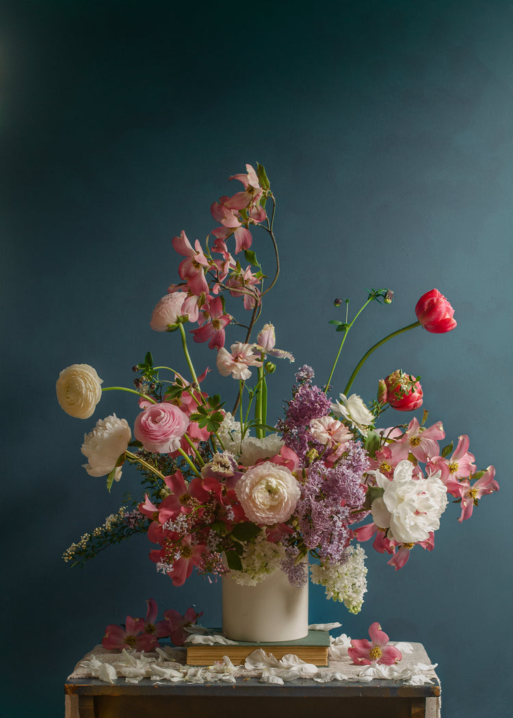 The Spring Ballet Poster showcases a lively floral arrangement in a white vase set against a dark teal background. The bouquet boasts pink, red, and white blooms such as ranunculus, peonies, and lilacs on a wooden table adorned with scattered petals.