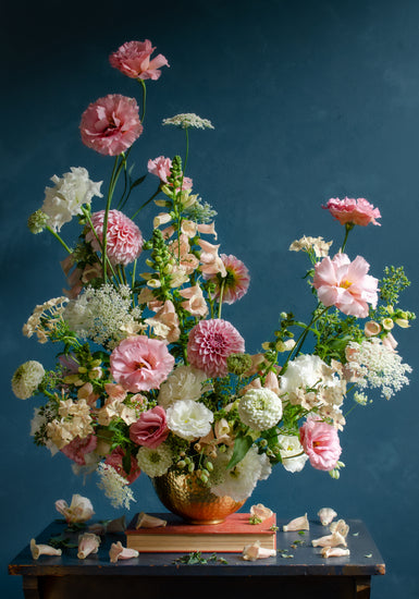 The Midsummer Poster showcases a floral arrangement with pink and white roses and peonies in a vase on a dark wooden table against a smooth, muted blue background, with a few petals scattered on the surface.