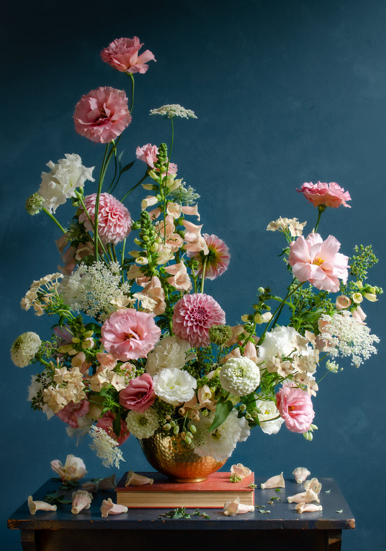 The Midsummer Poster showcases a floral arrangement with pink and white roses and peonies in a vase on a dark wooden table against a smooth, muted blue background, with a few petals scattered on the surface.