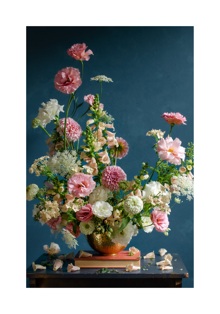 The Midsummer Poster showcases a vibrant floral arrangement in pink, white, and peach hues with green foliage in a gold vase on a small table. Its set against a dark blue backdrop with petals scattered around the base.