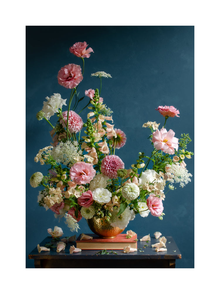 The Midsummer Poster features a lush arrangement of pink, white, and cream flowers, including carnations and dahlias in a bronze vase on a wooden table against a deep blue background with scattered petals enhancing the elegance and natural charm.