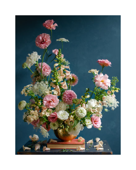 The Midsummer Poster showcases a vibrant floral arrangement of pink, white, and peach flowers with lush green stems and leaves in a textured vase. Set against a rich blue background, petals and sprigs are artistically scattered on the table below.
