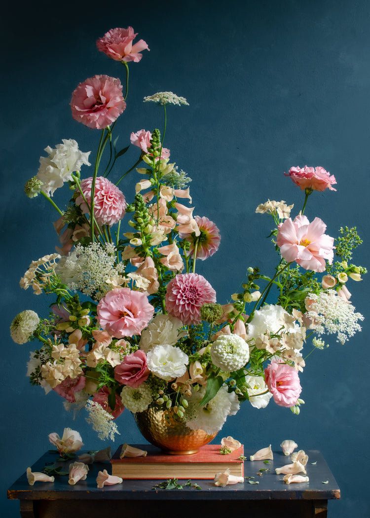 The Midsummer Poster showcases a vibrant floral arrangement with pink, white, and peach blossoms complemented by green foliage in a vase on a table against a dark blue background, with scattered petals adding elegance.