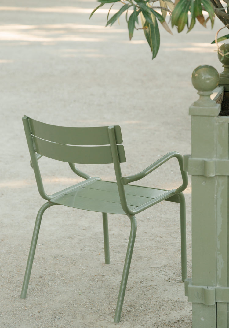 A serene, sunlit setting features a solitary green metal chair on a sandy path near a decorative post and leafy branch, casting soft shadows. This scene is beautifully captured in the Asseyez-vous Poster.