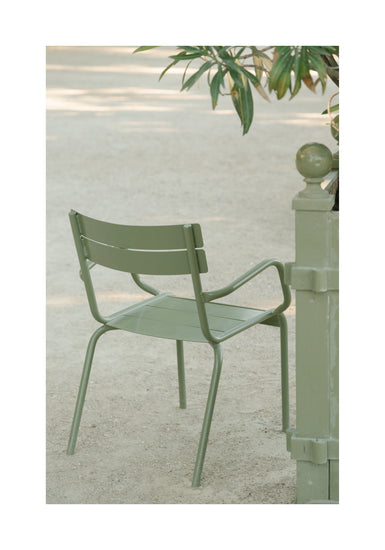 An Asseyez-vous Poster features a green metal chair on a sandy surface near a matching post, with green plants in the background conveying a tranquil outdoor atmosphere.