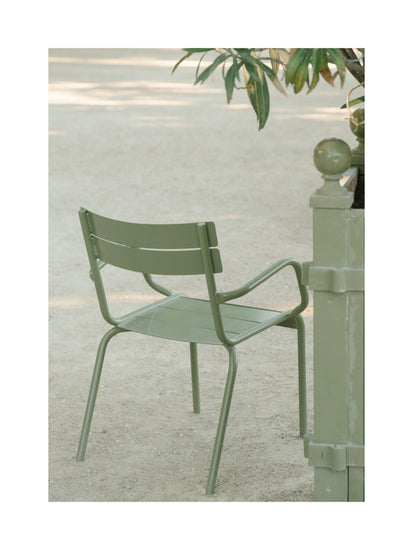 The Asseyez-vous Poster depicts a green metal chair with horizontal slats on sandy ground next to a cylindrical post with an orb. A leafy plant peeks above the post, setting a tranquil outdoor scene.