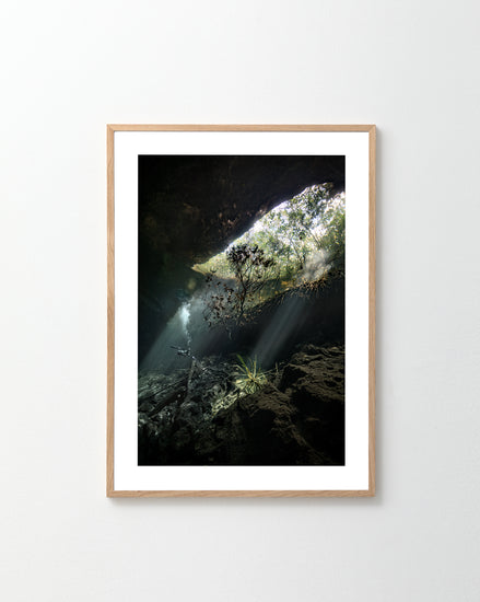 The Grotto Poster, a framed photograph of an underwater cave with sunlight filtering through the opening, creates a serene and mystical atmosphere. Rocks and plants add depth, while the light wood finish frame complements a white wall, inviting exploration without causing claustrophobia.