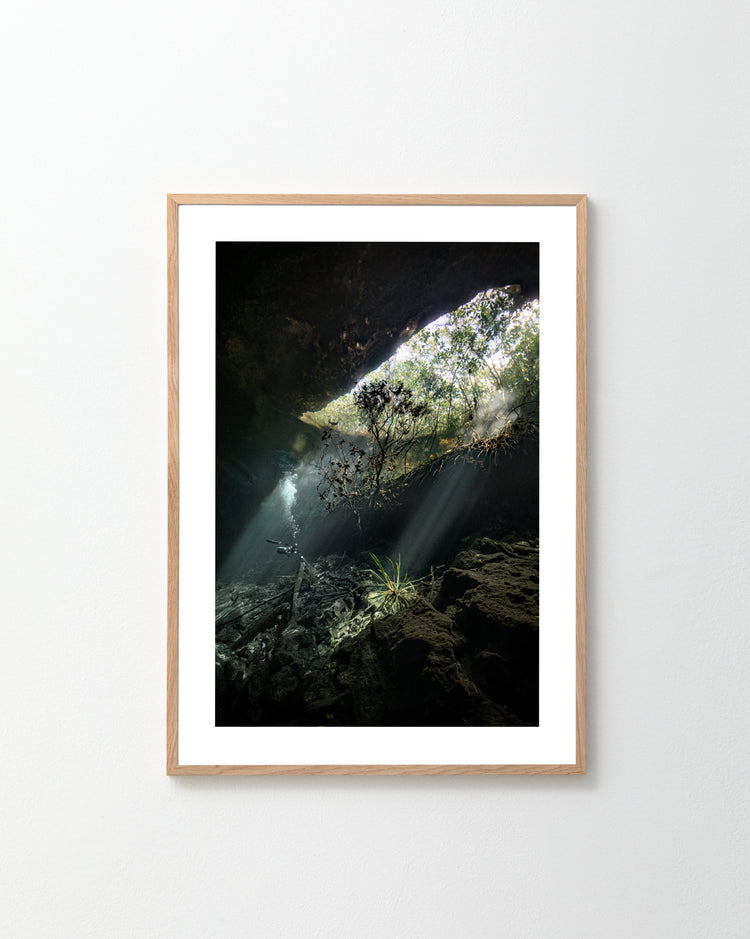 The Grotto Poster, a framed photograph of an underwater cave with sunlight filtering through the opening, creates a serene and mystical atmosphere. Rocks and plants add depth, while the light wood finish frame complements a white wall, inviting exploration without causing claustrophobia.