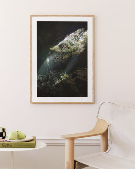 The Grotto Poster, showcasing sunlight through a cave over rocky surfaces and vegetation, enhances the rooms white wall. Beneath, a light wooden chair complements a round white table holding a green apple and a book.