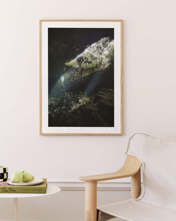 The Grotto Poster, showcasing sunlight through a cave over rocky surfaces and vegetation, enhances the rooms white wall. Beneath, a light wooden chair complements a round white table holding a green apple and a book.