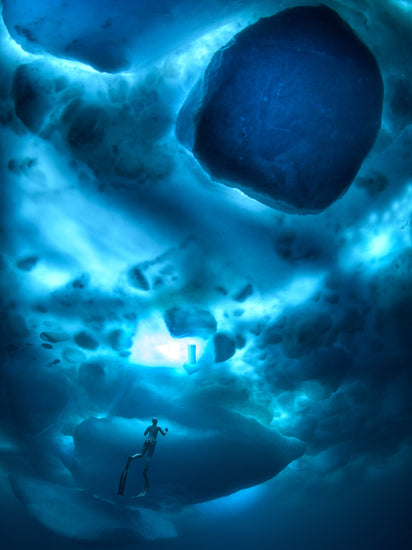 A diver swimming in the deep blue ocean underneath a surface made from ice.