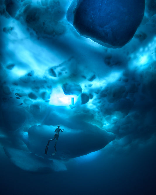 A diver swimming in the deep blue ocean underneath a surface made from ice.