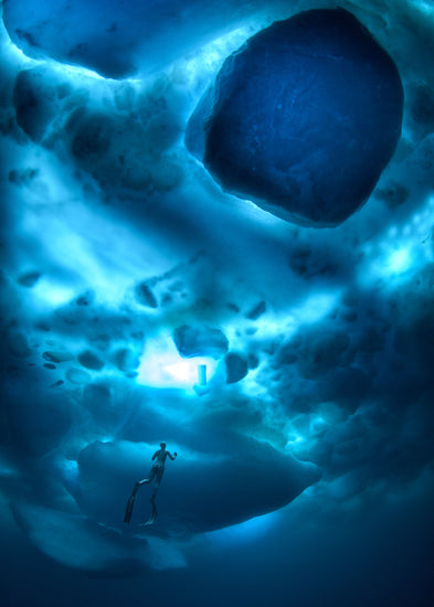 A diver swimming in the deep blue ocean underneath a surface made from ice.