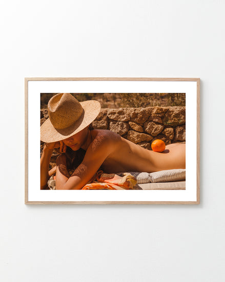 Naked woman on a sun bed wearing a hat and balancing an orange on her back.