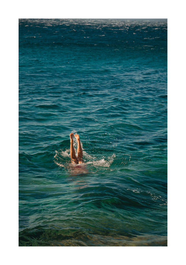 A person dives into the ocean during a morning swim, their legs just above the water. The sea reveals deep and light blue hues, with waves embracing them—a scene captured in the Morning Dip Poster reminiscent of a Lavinia Cernau wall-hanging.