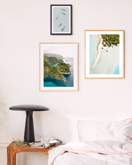 A cozy bedroom corner showcases a wooden side table with a sleek black lamp and a glass of water. Three framed aerial landscape photos, including the Piña Colada as a Place Poster featuring sharks, lush islands, and a beach shoreline, adorn the white wall. A tidy white bed is partially visible.