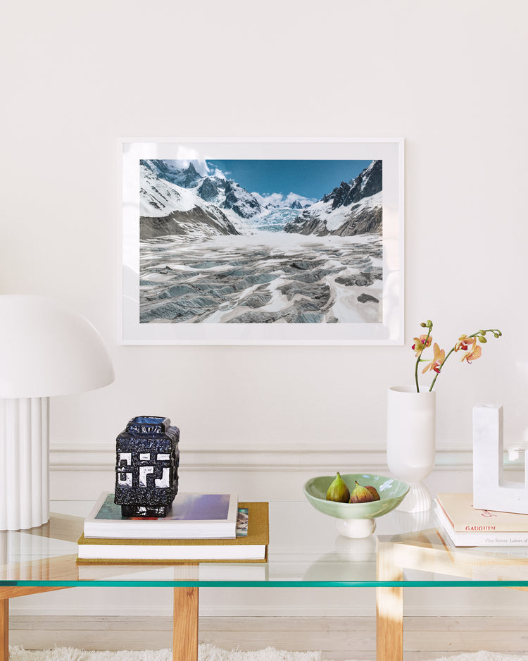 A modern living room mirrors nature, featuring a glass table with books, a decorative container, a green plate with figs, and an orchid in a vase. A white lamp stands left of the Greetings From Planet Earth I poster of snowy mountains reminiscent of Sir David Attenboroughs explorations.