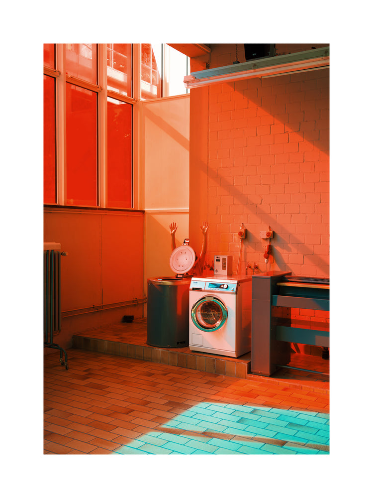In a stylish laundromat-inspired room with orange and teal hues, a washing machine and sink rest against taupe walls. Sunlight streams through large windows, highlighting a mannequin torso with raised arms, capturing the artistry of the Laundry Day Poster by Delfina Carmona.