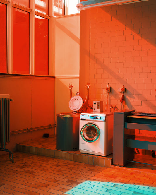 A laundry room, like a scene from the Laundry Day Poster, glows in red-orange light. A washing machine stands next to a trash can with mannequin arms in a basket on top, and large windows above a radiator cast shadows on the tiled floors laundromat-like atmosphere.