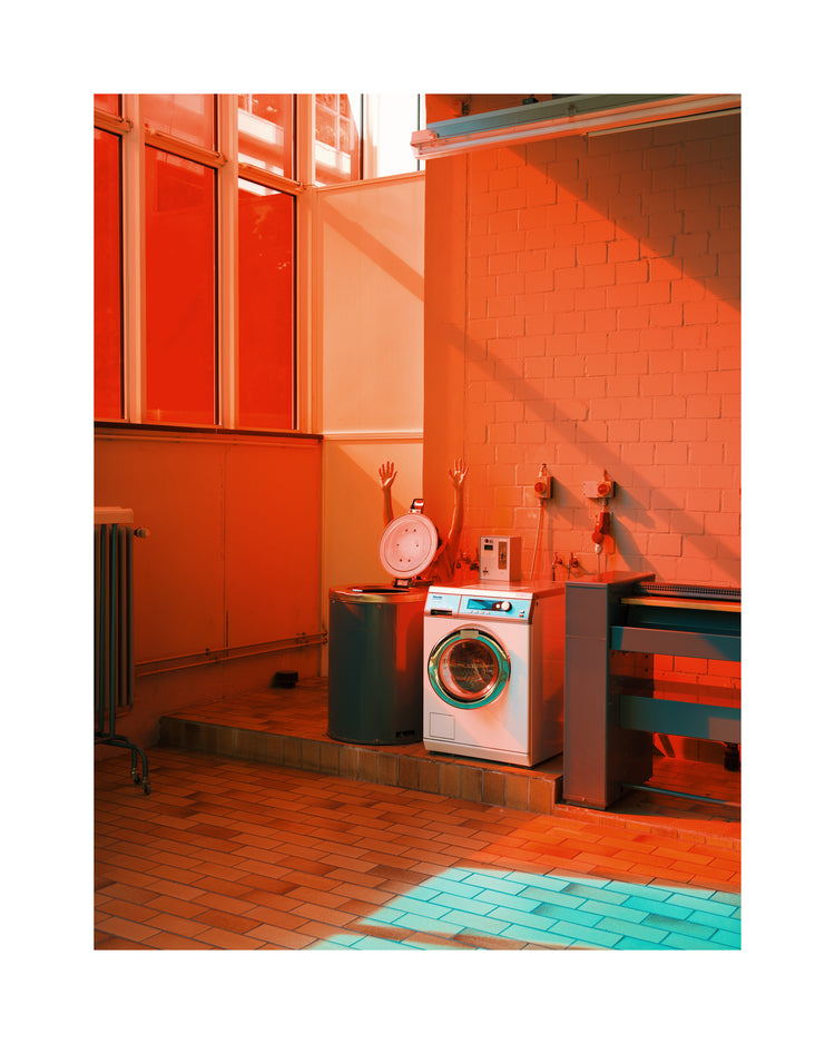 Brightly lit with orange and teal hues, a laundromat features a washing machine, a large industrial sink, and a weighing scale. Sunlight through tall windows casts colorful shadows on the brick wall and tiled floor, evoking the charm of the Laundry Day Poster.