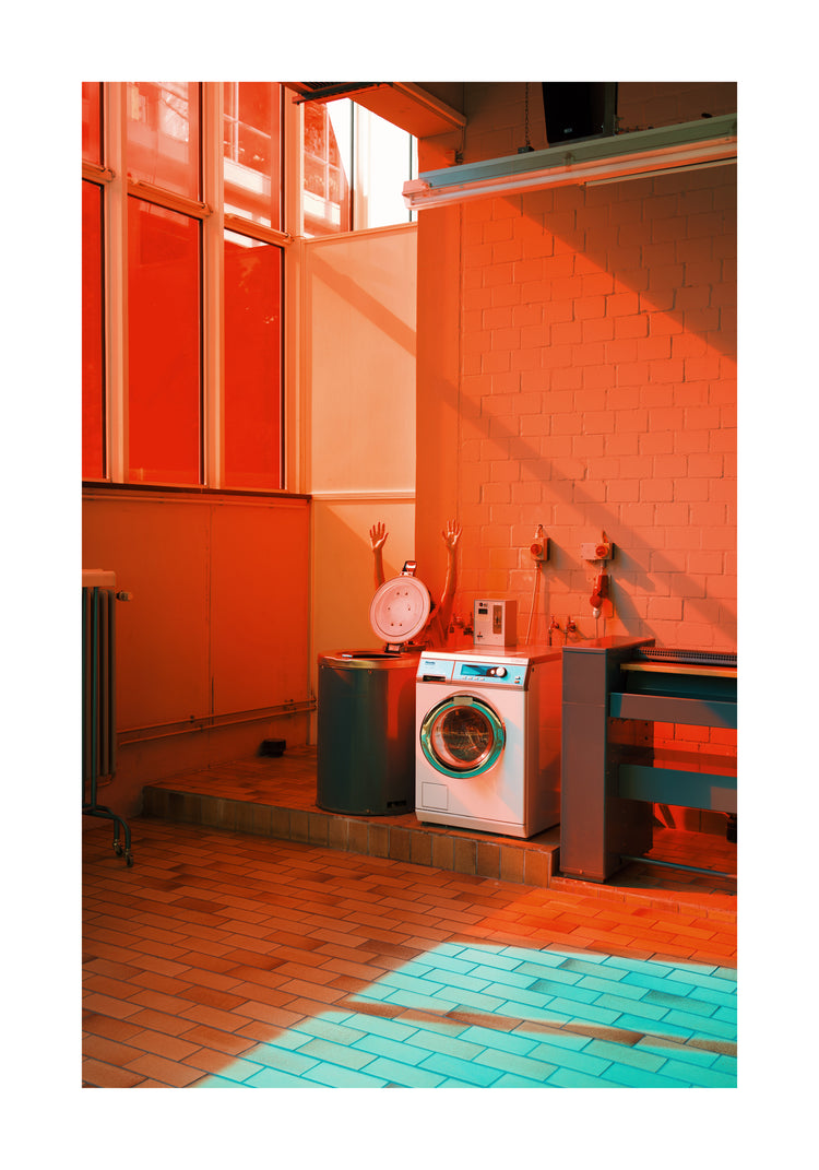 A dimly lit laundromat with red and teal lighting showcases a washing machine, a mannequin arm-laden basin, and exposed pipes against a brick wall. Laundry Day posters by Delfina Carmona beautify the space as large windows cast colored shadows over the tiled floor.