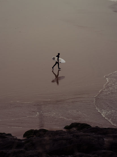 Surfer in a black wetsuit carrying a small board heading out to sea.
