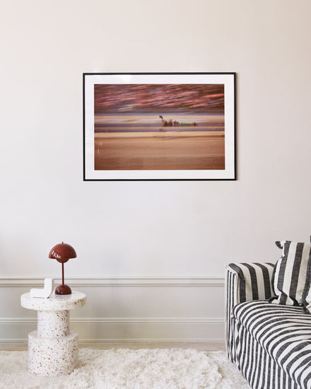 The Limitless Poster, a framed abstract beach scene, hangs on a beige wall above a modern round speckled table with a brown lamp. Nearby, a gray and white striped couch is enhanced by two accent pillows.