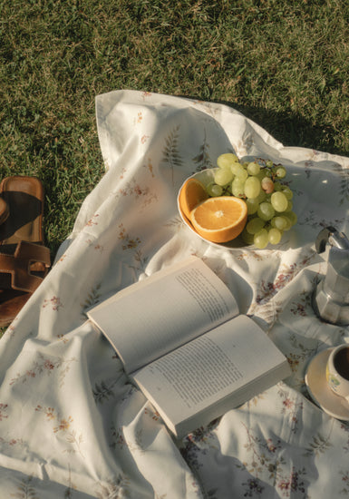 A Tuscan Summer Poster depicts a serene picnic with an open book on a floral blanket, a bowl of grapes, an orange half, a small coffee pot and cup, and brown sandals on lush grass, capturing leisurely moments enriched by natures simple pleasures.