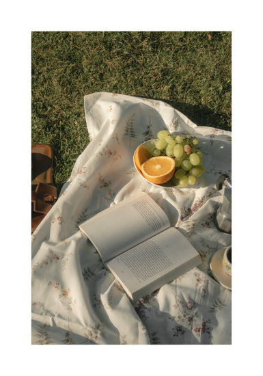The Tuscan Summer Poster captures an idyllic picnic scene with an open book on a floral blanket, a bowl of green grapes, a halved orange, and a cup nearby. A partially visible brown leather bag suggests an afternoon of serene leisure.