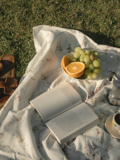 The Tuscan Summer Poster depicts a floral picnic blanket on grass, featuring an open book, a bowl of green grapes with orange halves, sandals, a stovetop coffee maker, and a cup. Sunlight gently highlights this serene scene, perfect for escaping into natures embrace.