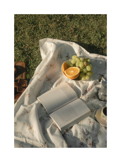 The Tuscan Summer Poster depicts a vivid picnic scene on lush grass with a floral blanket featuring an open book, green grapes and orange slices, alongside sandals and a steaming coffee cup and pot. Soft lighting enhances the warm, relaxed atmosphere of this idyllic setting.