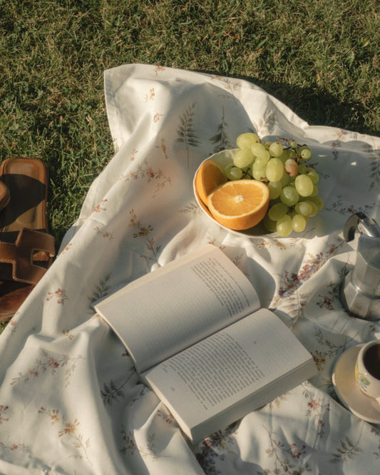 The Tuscan Summer Poster captures an idyllic outdoor picnic scene, featuring a floral blanket on grass with an open book, a plate of sliced oranges and grapes, a coffee maker, and a cup. Nearby lie sandals, completing the serene escape in natures embrace.
