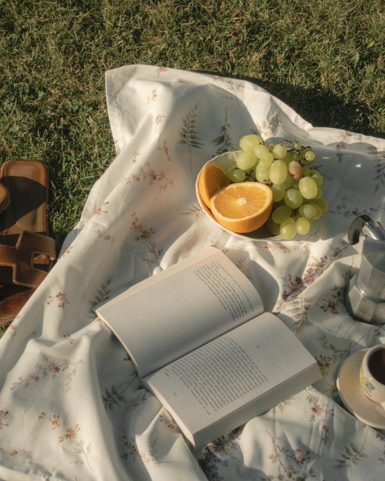 The Tuscan Summer Poster captures an idyllic outdoor picnic scene, featuring a floral blanket on grass with an open book, a plate of sliced oranges and grapes, a coffee maker, and a cup. Nearby lie sandals, completing the serene escape in natures embrace.