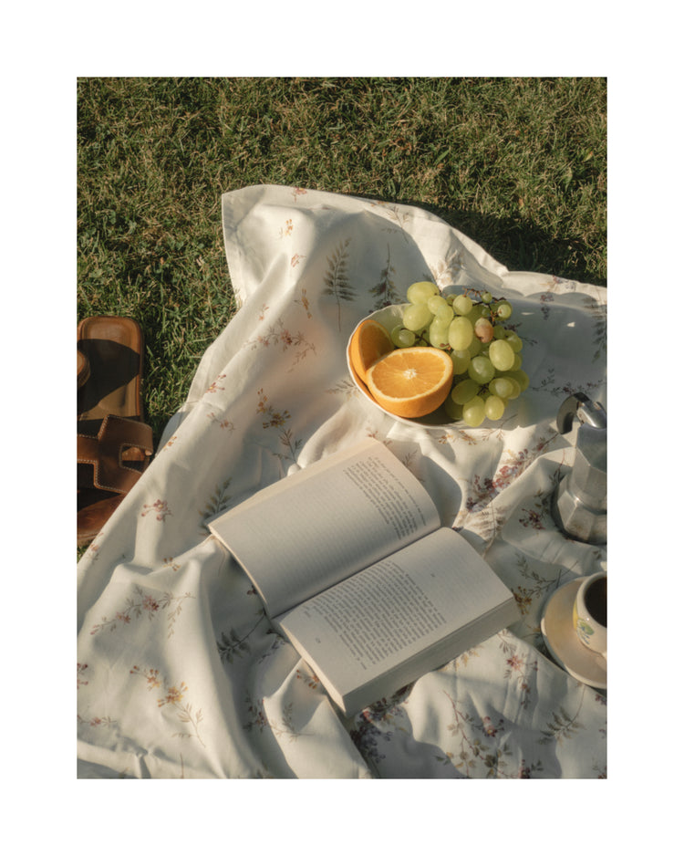 In the Tuscan Summer Poster, a picnic scene features a floral blanket with an open book and enticing grapes, a halved orange in a bowl, and an aromatic cup of coffee. A leather bag rests on the grassy ground.