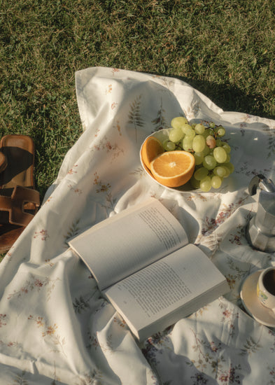 The Tuscan Summer Poster captures an enchanting picnic escape with a floral blanket on a grassy field. Juicy grapes, half an orange, sandals, and aromatic coffee create a serene ambiance. A metallic coffee pot gleams under the suns soft shadows, completing this idyllic retreat.