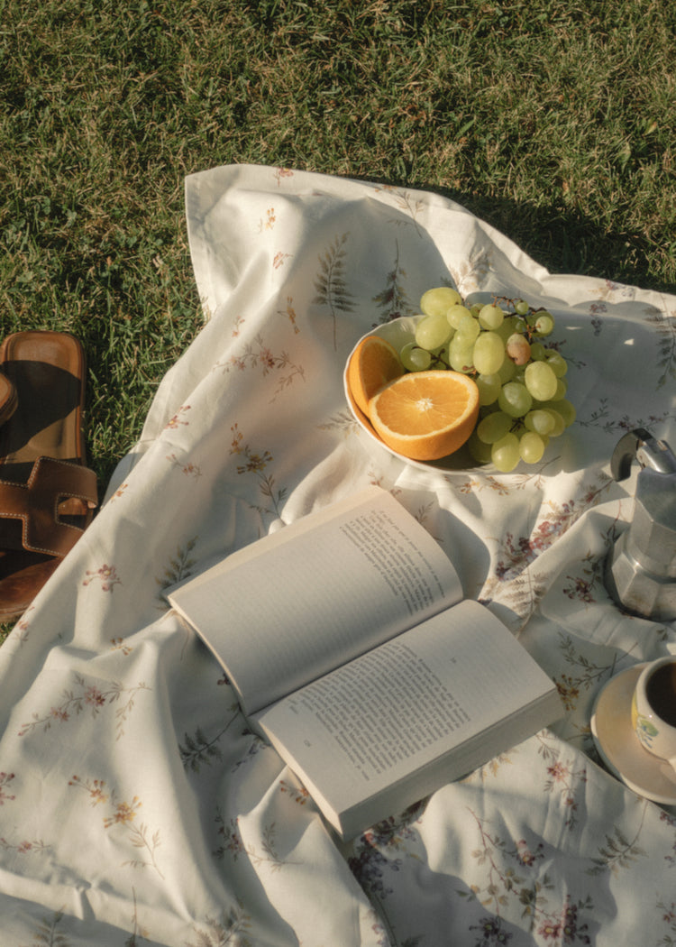 The Tuscan Summer Poster captures an enchanting picnic escape with a floral blanket on a grassy field. Juicy grapes, half an orange, sandals, and aromatic coffee create a serene ambiance. A metallic coffee pot gleams under the suns soft shadows, completing this idyllic retreat.