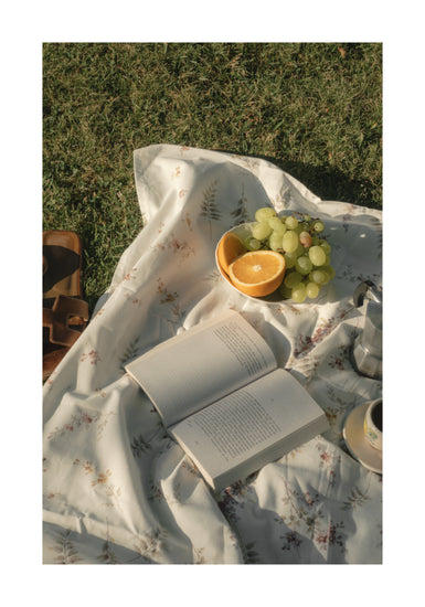 The Tuscan Summer Poster depicts a picnic scene on grass featuring a floral blanket with an open book, a bowl of green grapes, and half an orange. A brown leather bag and ceramic cup are partially visible as sunlight casts soft shadows.