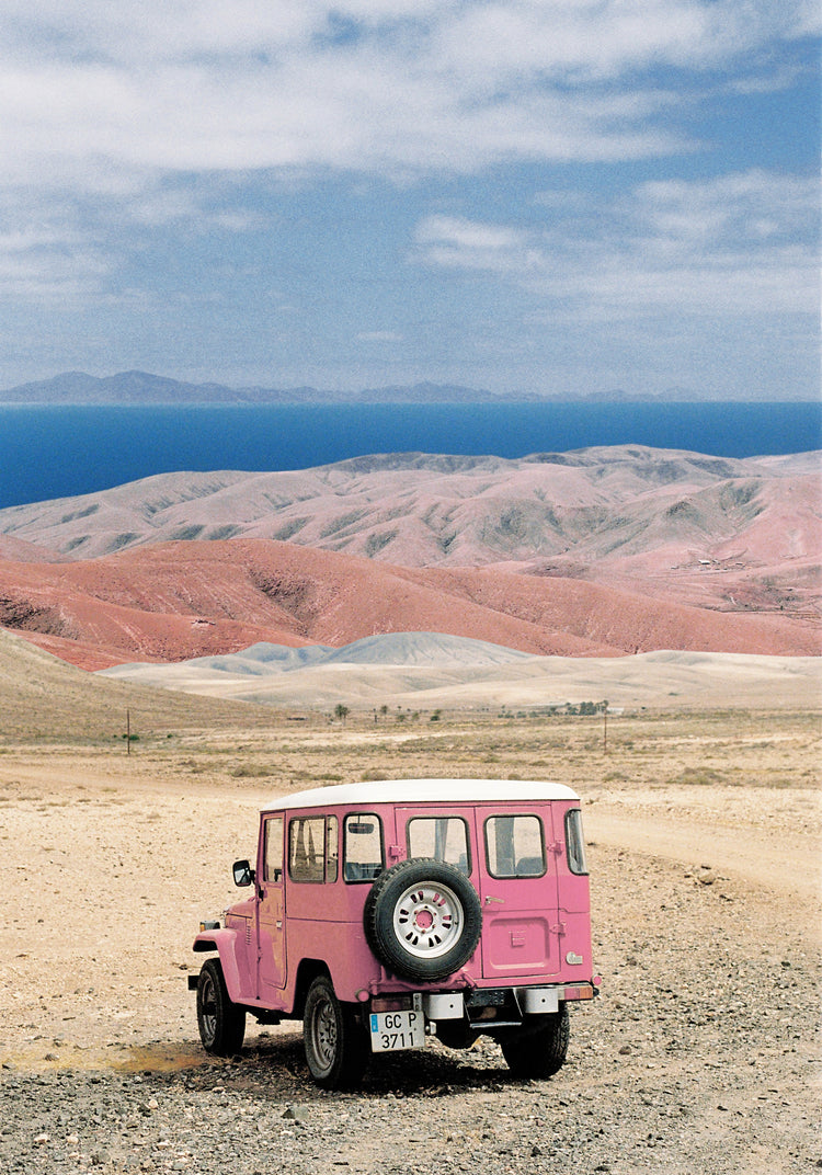 Pink jeep standing in a desert with pink sand dunes and an ocean in the horizon.