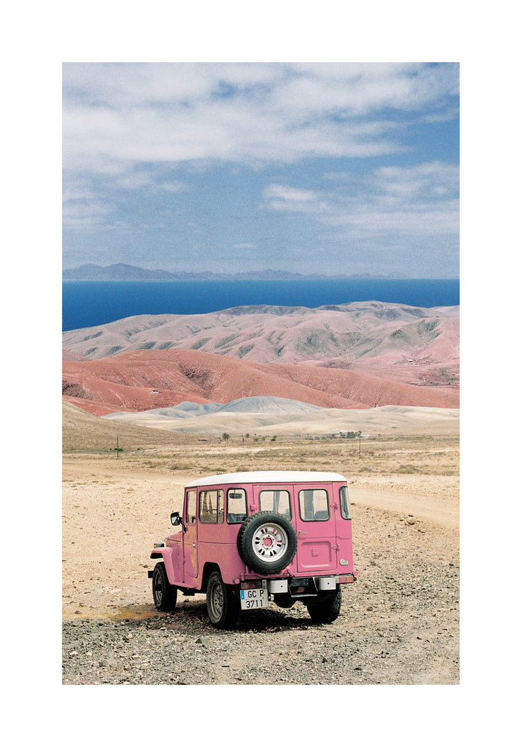 Pink jeep standing in a desert with pink sand dunes and an ocean in the horizon.