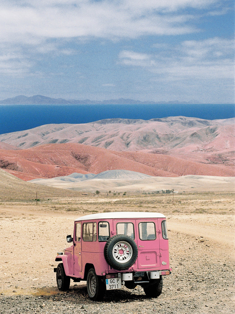 Pink jeep standing in a desert with pink sand dunes and an ocean in the horizon.