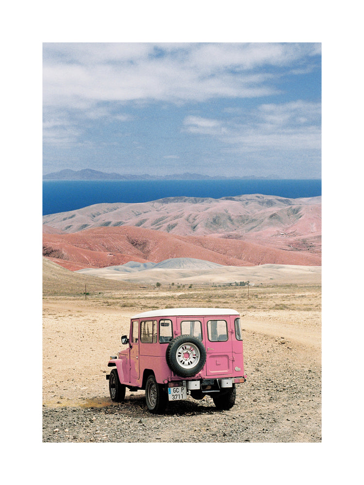 Pink jeep standing in a desert with pink sand dunes and an ocean in the horizon.