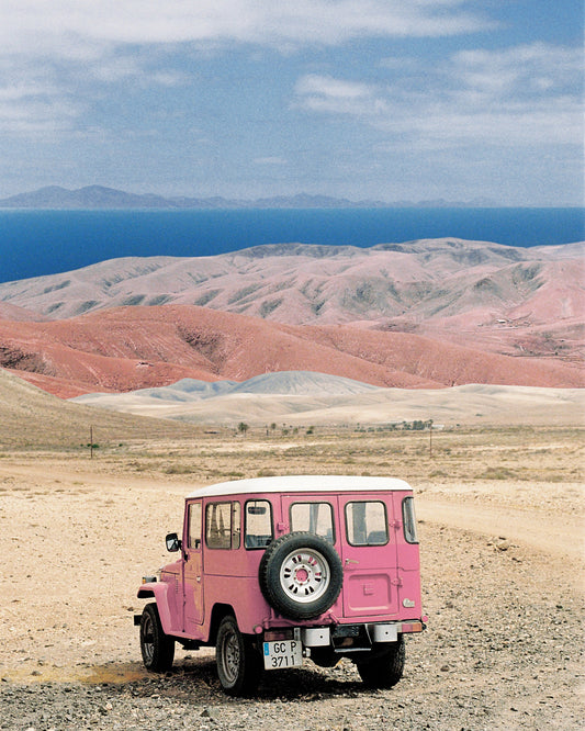 Pink jeep standing in a desert with pink sand dunes and an ocean in the horizon.