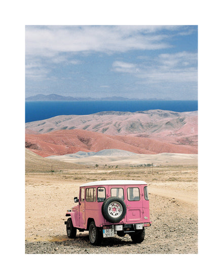 Pink jeep standing in a desert with pink sand dunes and an ocean in the horizon.