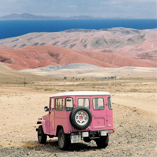Pink jeep standing in a desert with pink sand dunes and an ocean in the horizon.
