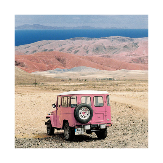 Pink jeep standing in a desert with pink sand dunes and an ocean in the horizon.