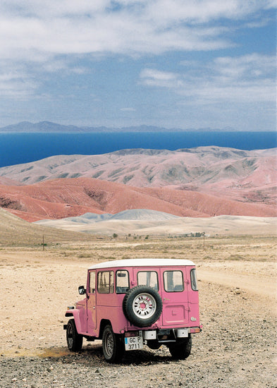 Pink jeep standing in a desert with pink sand dunes and an ocean in the horizon.