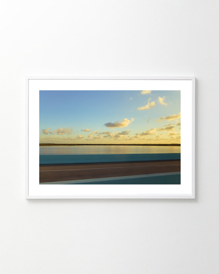 The Water Split Poster showcases a serene landscape with a body of water divided by land, resembling Moses parting the sea, under a vast sky dotted with fluffy clouds and bathed in warm sunlight.