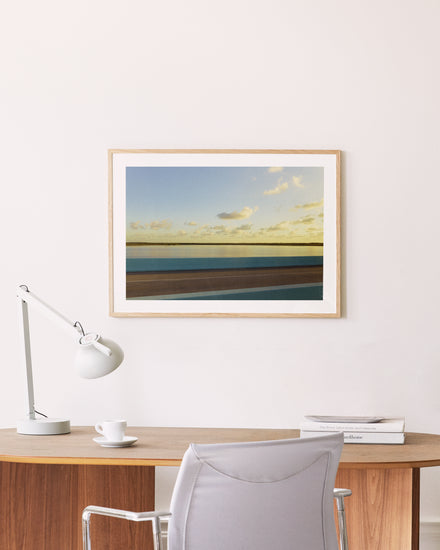 A minimalist wooden desk with a white lamp, cup, and books sits beside the road to creativity. In front is a light gray chair. Gracefully hanging above on the cream-colored wall is a wooden-framed Water Split Poster depicting clouds and water.