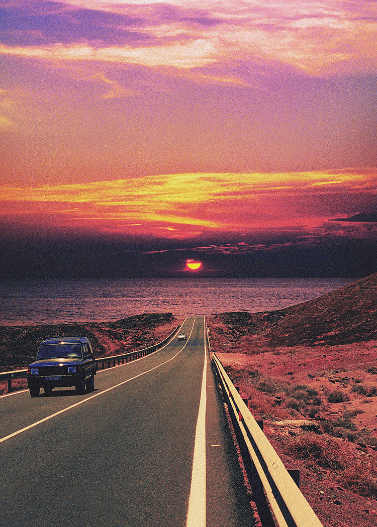 Road winding its way towards the ocean where a sun is setting on the horizon.