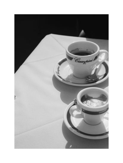 This Ciampini Poster showcases a vintage Rome vibe with black and white photography: two elegantly poised coffee cups on saucers, complete with spoons, sit on a draped tablecloth. The larger cup holds coffee and foam; the smaller, espresso. Sunlight casts soft shadows.